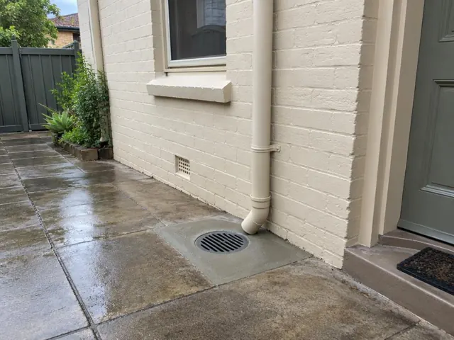 Outdoor drain grate beside a brick house wall and paved side path in South Granville